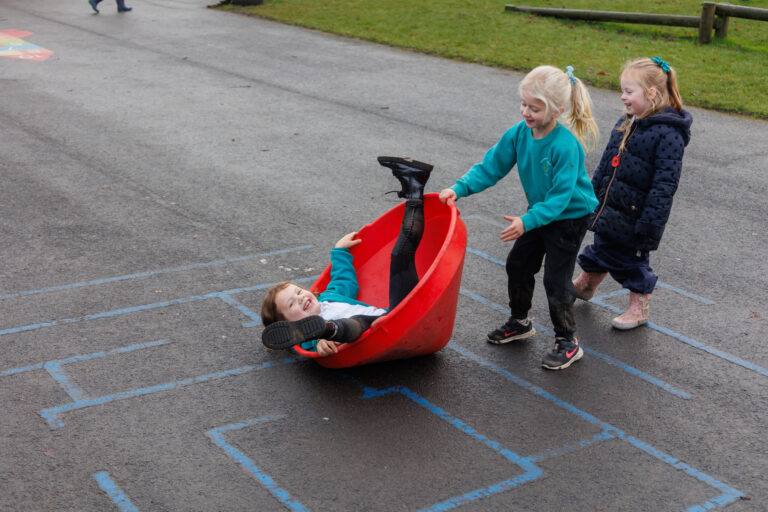 A group of children playing outside on tarmac, with one kid lying in a red shaped cone.