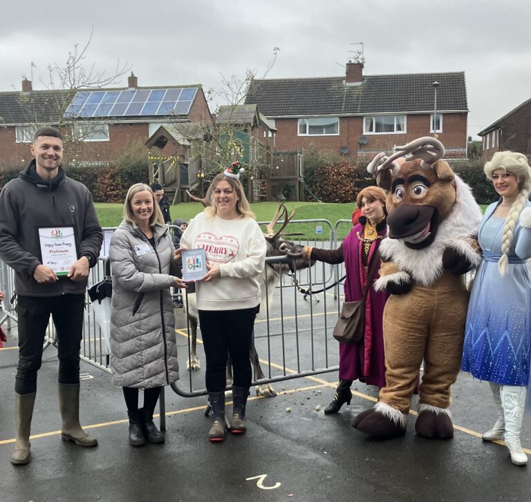 People standing outside in a playground, holding certificates. Some on the right are dressed in fancy dress and standing next to a reindeer mascot.