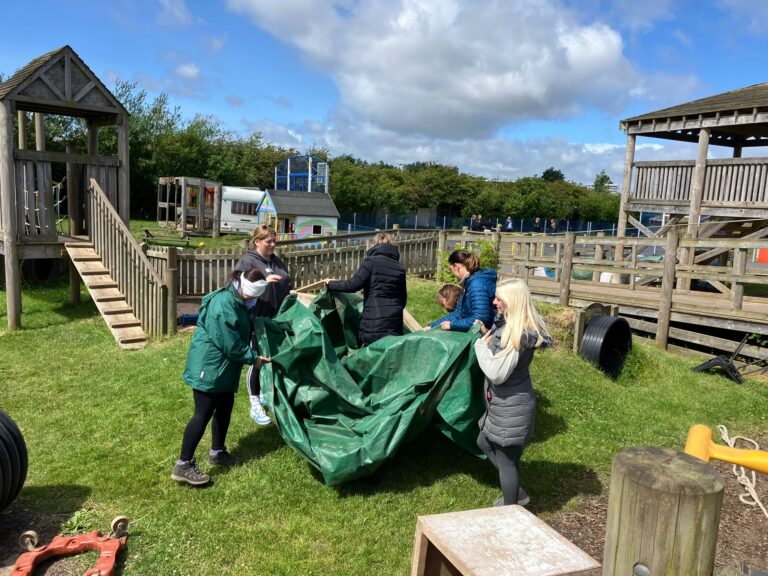 An image of a group of people in a park, holding a tarpaulin. It is a sunny day.