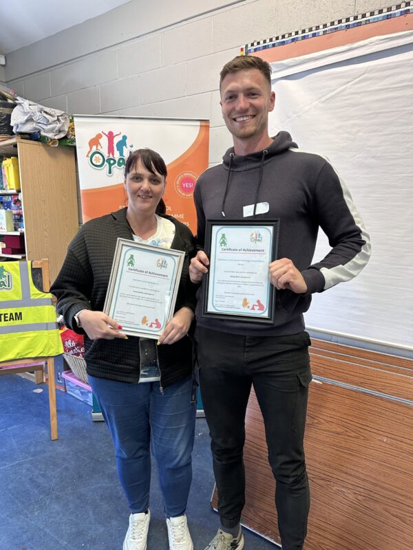 A woman and a man standing next to each other, each holding an award in a classroom.
