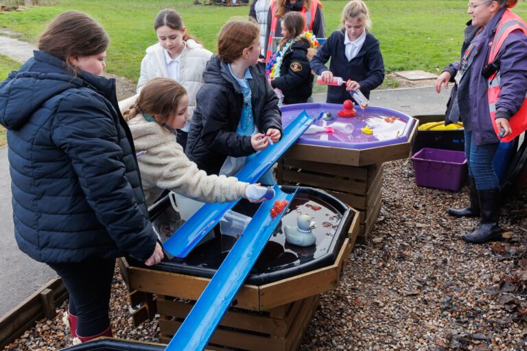 Six kids playing with toys outdoors in a garden, with a woman standing next to them wearing an orange high visibility jacket.