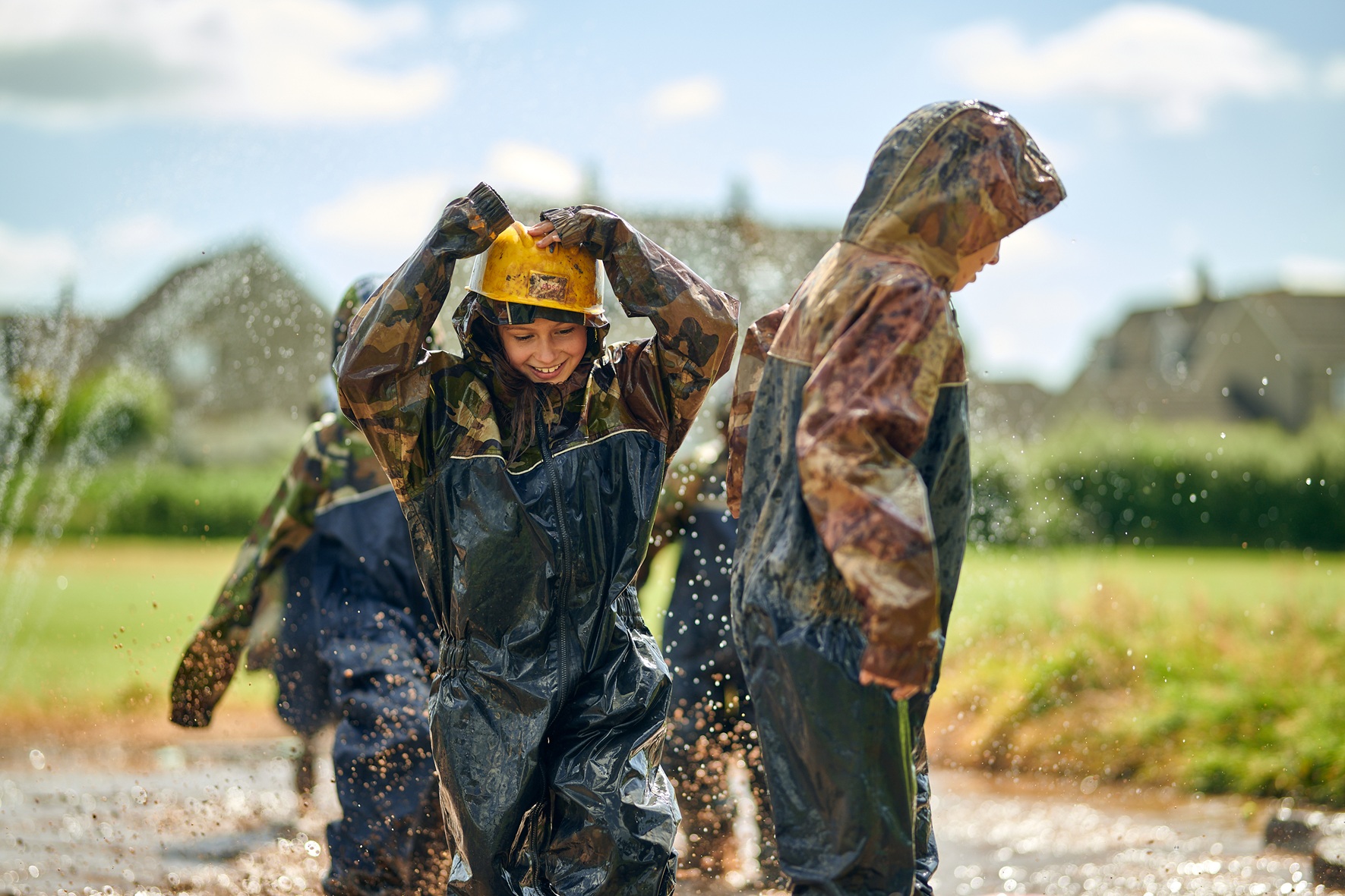 Three children playing in a mud pit at school. They are wearing head-to-toe waterproofs.