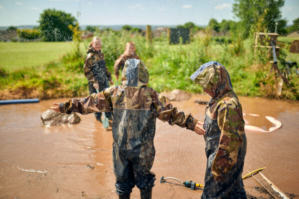 A group of children play outdoors at school in a big muddy puddle. They are wearing head-to-toe waterproofs and splashing around.